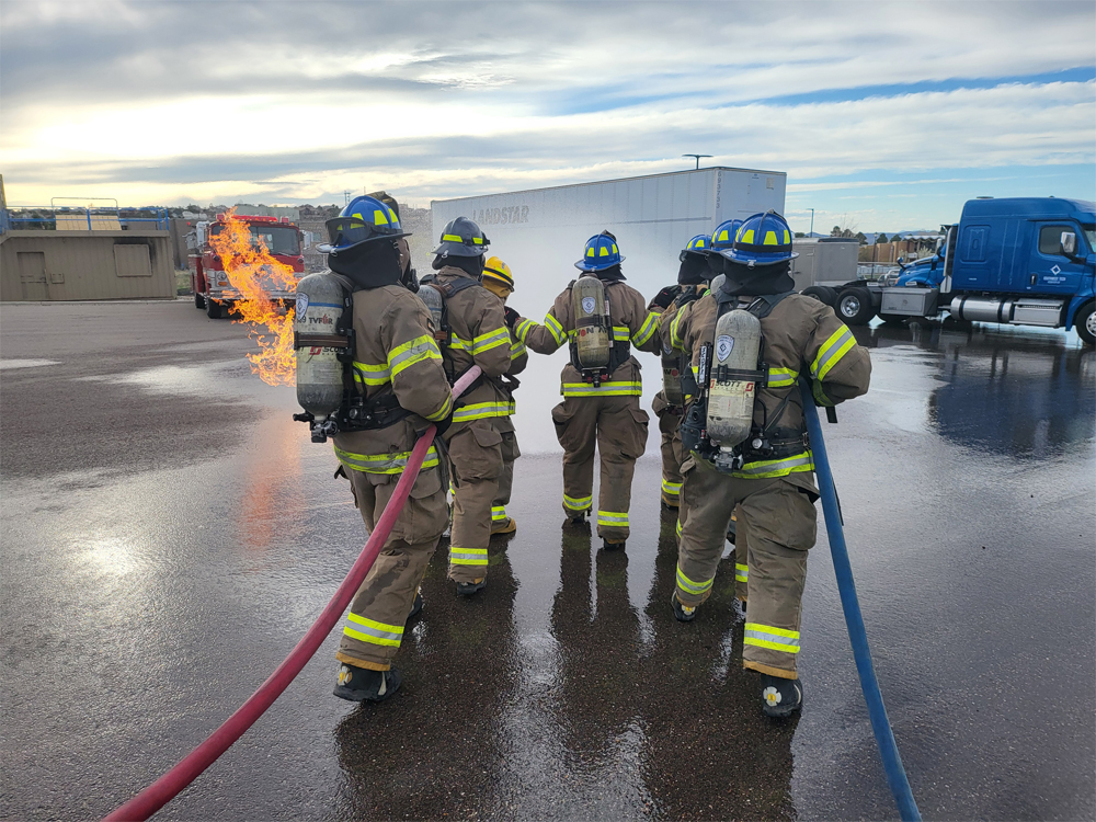 Firefighters in turnout gear spray hoses during outdoor live fire training.