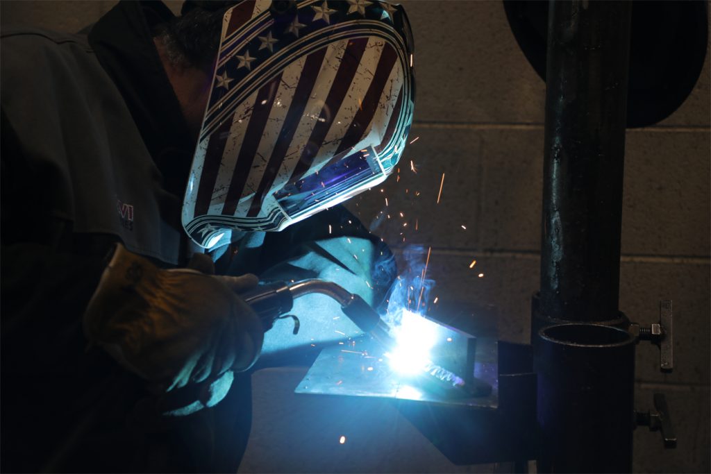 Welder in an American flag helmet sparks metal at a workbench.