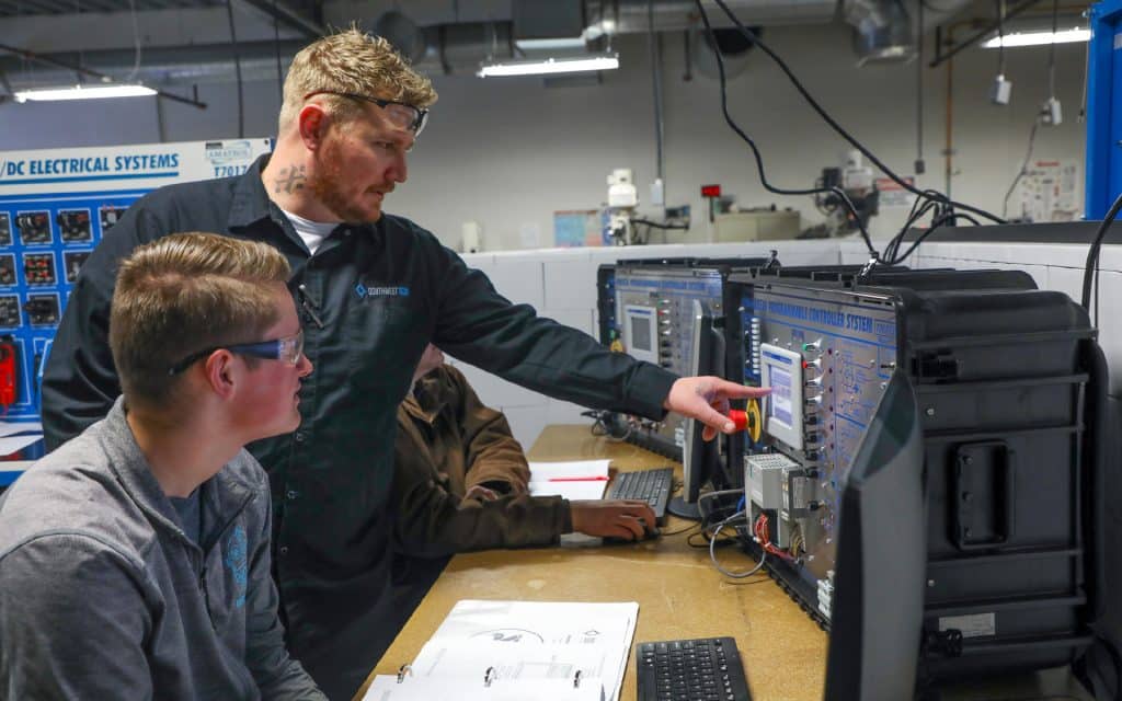 Instructor in safety glasses points to a programmable controller for a student.
