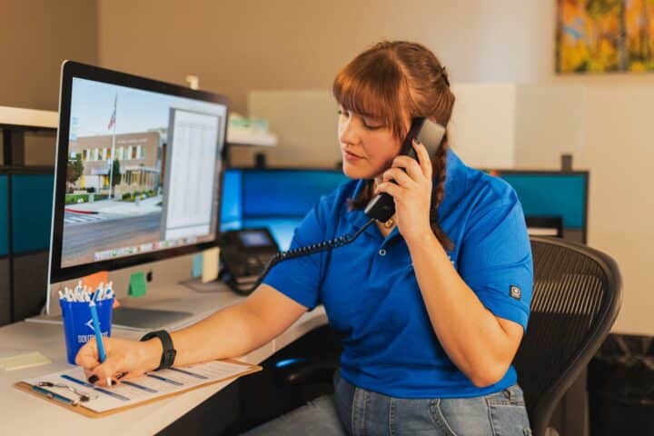 Staff member in a blue polo takes a phone call while writing at a desk.