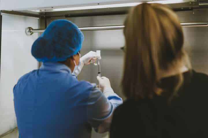 Student in scrubs and a hairnet draws from a vial inside a sterile hood.