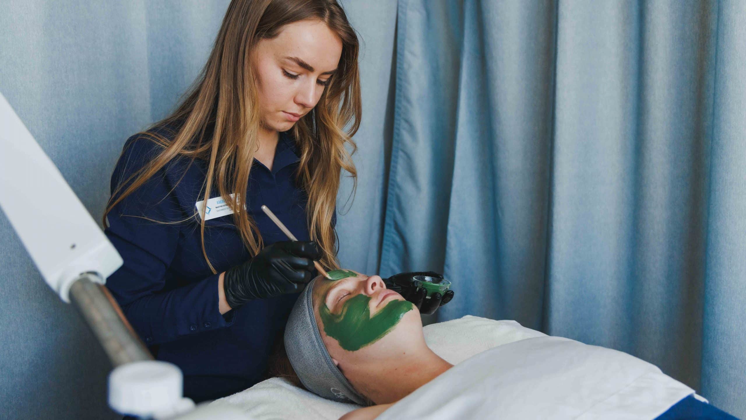 Esthetics student applies a green facial mask to a client on a treatment bed.