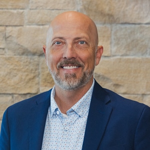 Brennan Wood in a navy blazer and dotted shirt against a stone wall.