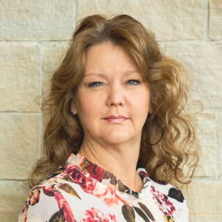 Patricia Weller in a floral blouse and hoop earrings against a stone wall.